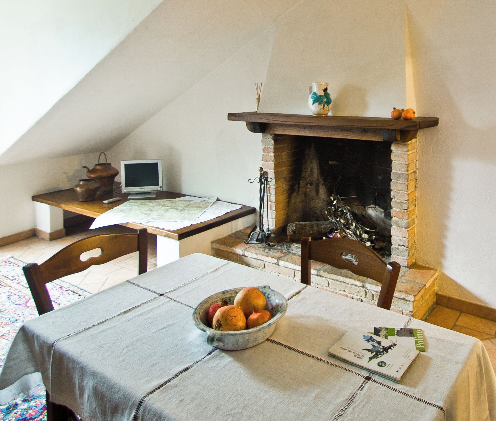 Une pièce chaleureuse avec une cheminée, une table à manger ornée d’un panier de fruits et un bureau avec un ordinateur et une lampe.