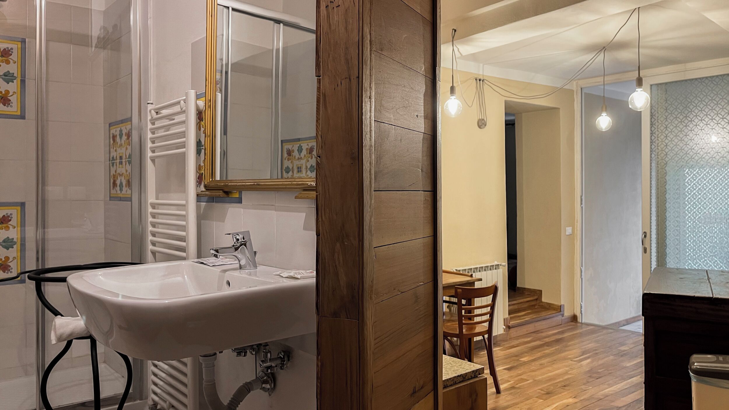 A bathroom with a white sink, a mirror, and a towel rack. There is a wooden door leading to a dining area with a table and chairs.