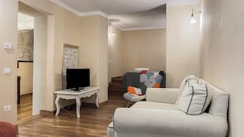 Un salón con una televisión de pantalla plana sobre una mesa blanca, un sofá color beige y una escalera que conduce a otra habitación.