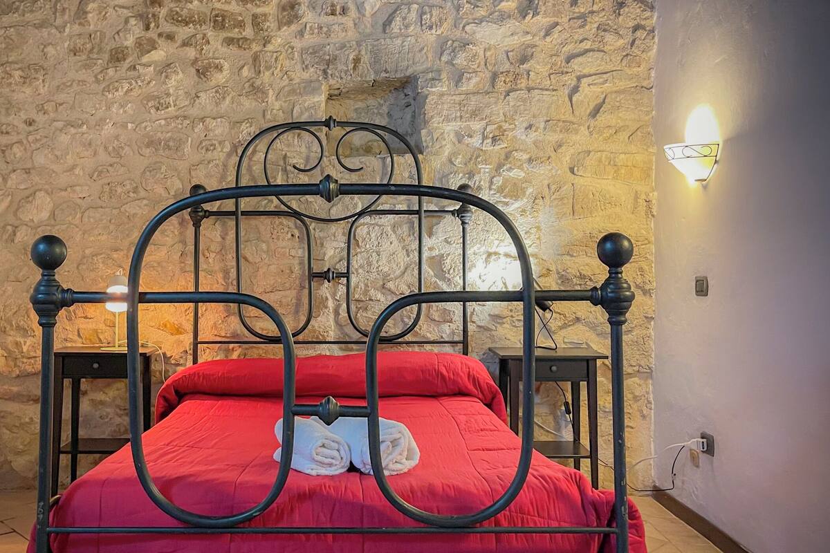 A bed with a metal frame and red bedding in a room with stone walls and a radiator.