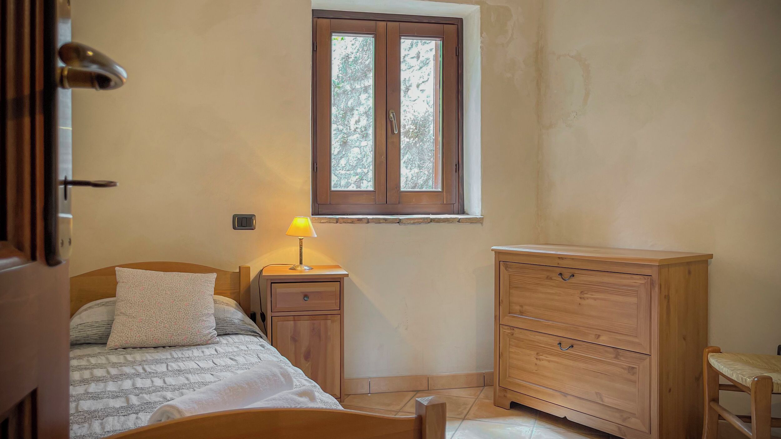 A bedroom with a bed, a nightstand with a lamp, a wooden dresser, and a window with a view of trees.