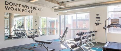 Une salle de sport équipée de matériel d’exercice, avec une citation motivante sur le mur et de grandes fenêtres.