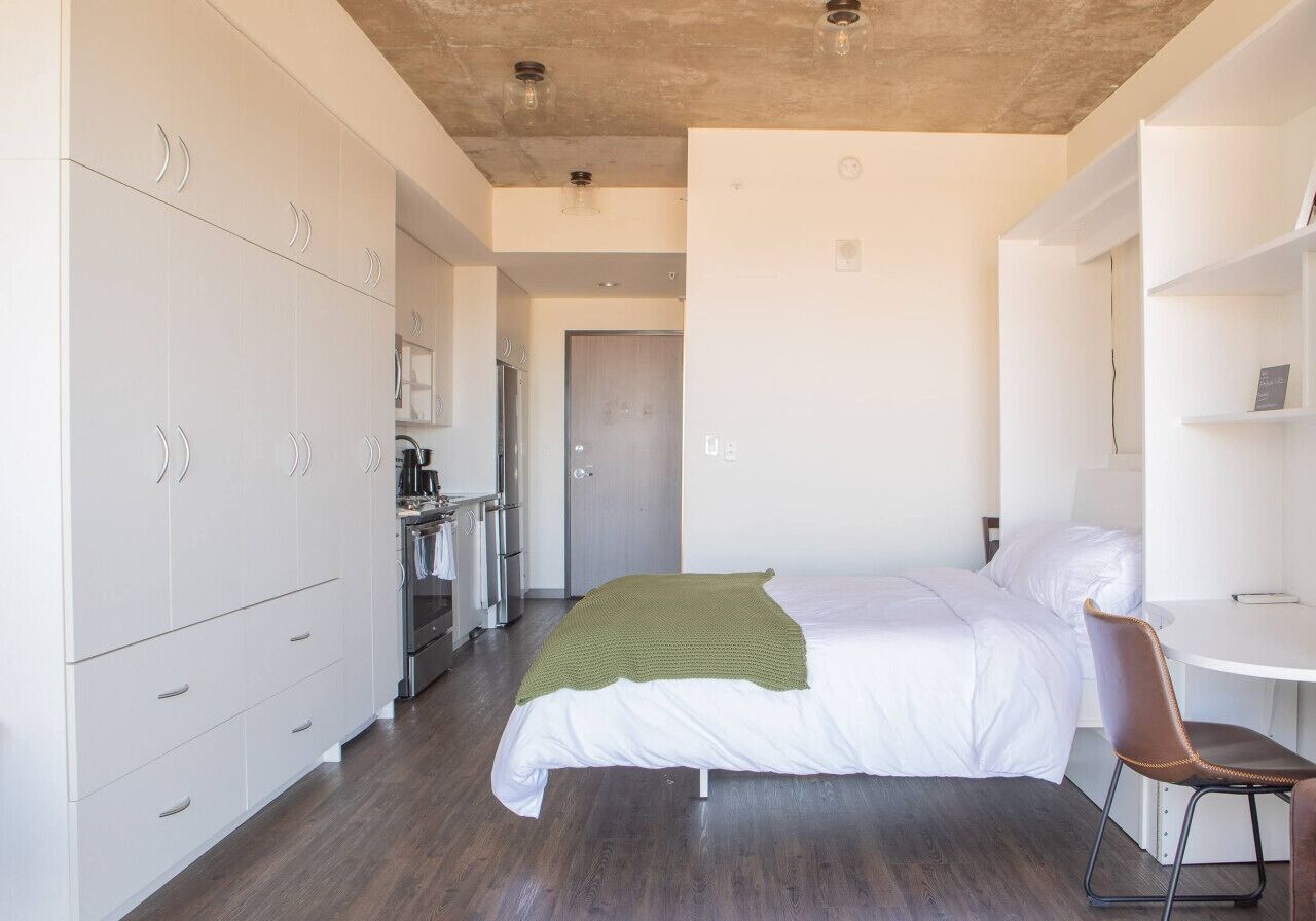 A modern hotel room with a large bed, a desk, and a kitchenette.