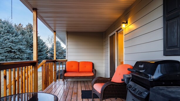 Eine überdachte Veranda mit Holzterrasse, einem Korbsessel mit orangenen Kissen, einem Glastisch und einem Grill.