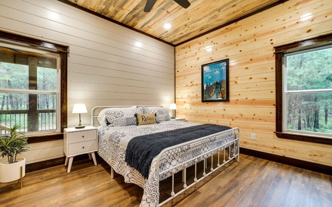 A bedroom with a wooden ceiling, a bed with patterned bedding, two bedside tables, and a large window.