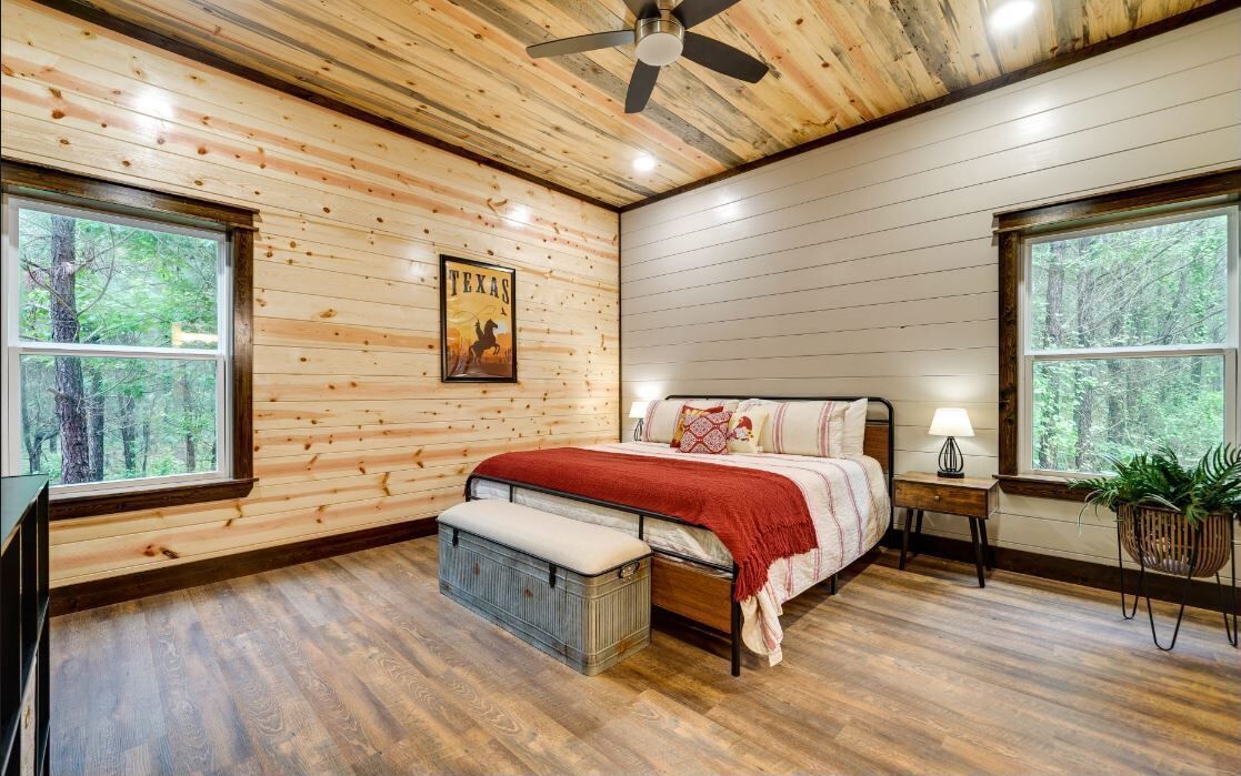 A bedroom with wooden walls, a bed with a red blanket, a nightstand, a lamp, and a window with a view of trees.