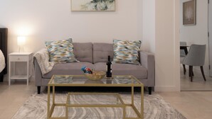 A living room with a sofa, a glass coffee table, a lamp, and a painting on the wall. - Large, Newly Built 1B Studio w/ Patio, W&D (Naples)