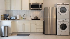 A compact kitchen with white cabinets, a stainless steel microwave, a built-in oven, a washing machine, and a trash bin. - Large, Newly Built 1B Studio w/ Patio, W&D (Naples)