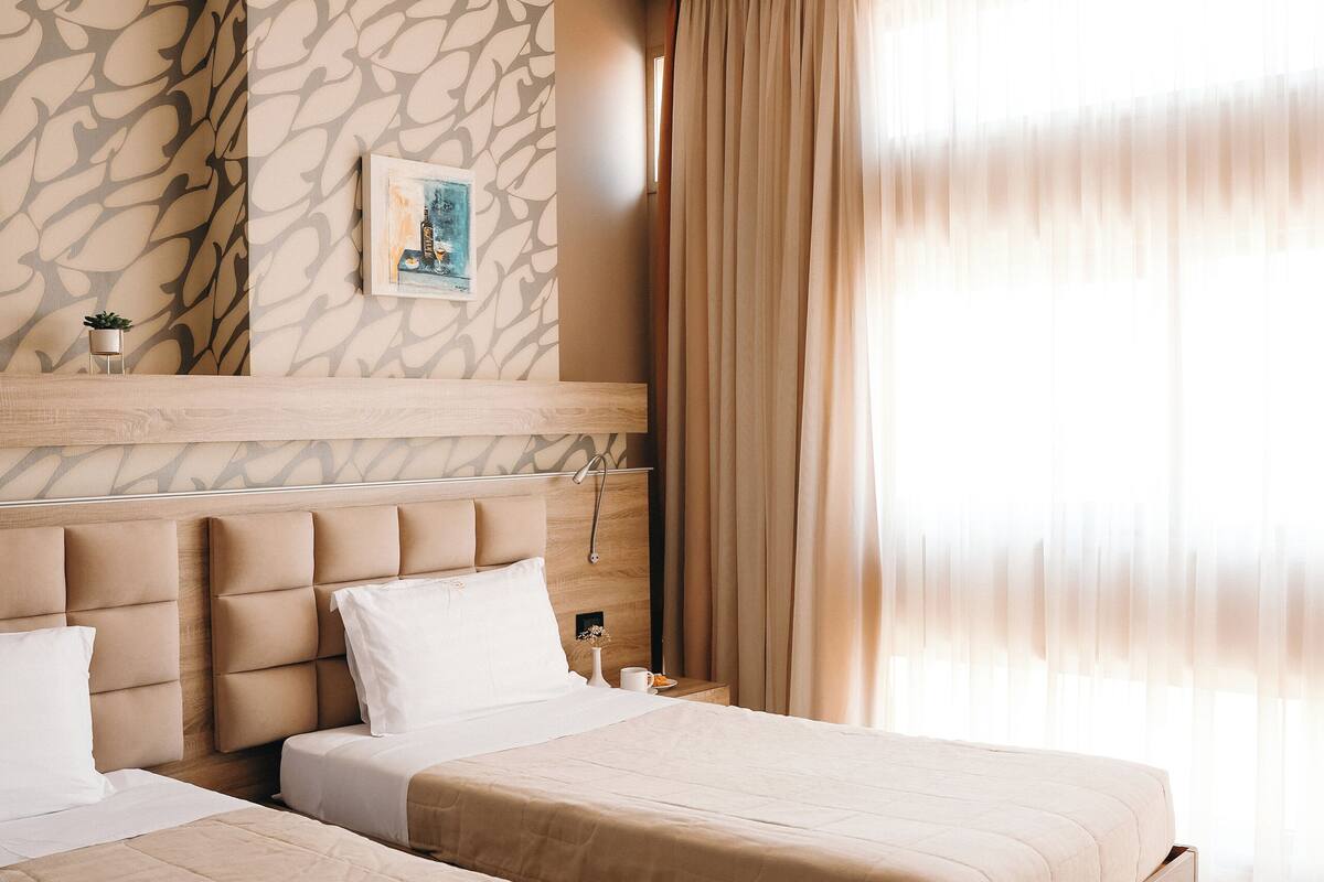 A hotel room with two beds, a stone-patterned wall, a framed picture, and a large window with curtains.