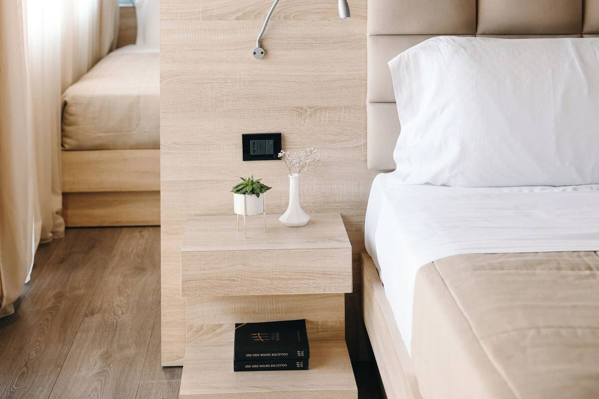 A modern hotel room with a wooden headboard, a bedside table with a small plant, a framed artwork above the bed, and a control panel on the wall.