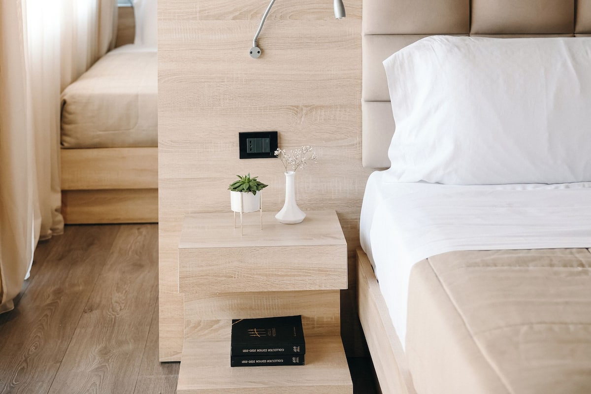 A modern hotel room with a wooden headboard, a bedside table with a small plant, a framed artwork above the bed, and a control panel on the wall.