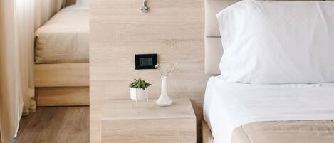 A modern hotel room with a wooden headboard, a bedside table with a small plant, a framed artwork above the bed, and a control panel on the wall.