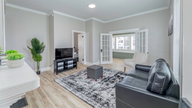 A modern living room with a black leather sofa, a grey ottoman, a black and white rug, a TV, and a plant.