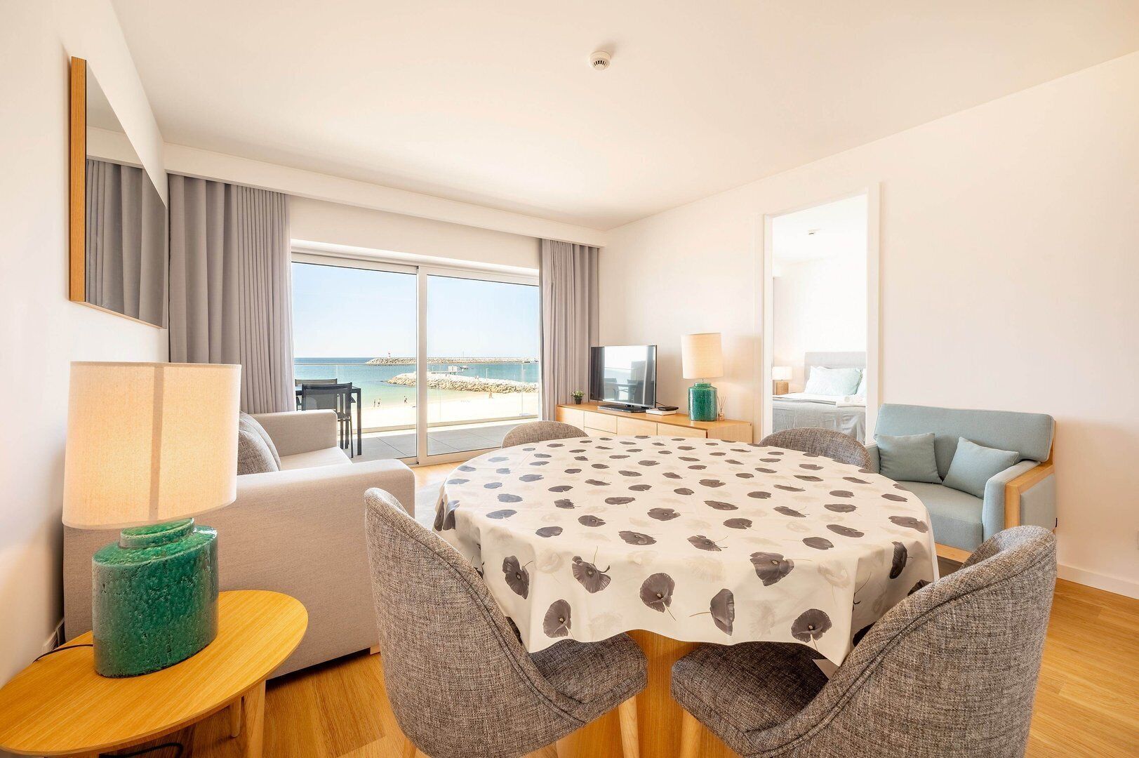 Habitació d'hotel amb un gran finestral que ofereix vistes a la platja, zona de menjador amb taula i cadires, sofà i televisió sobre un suport.