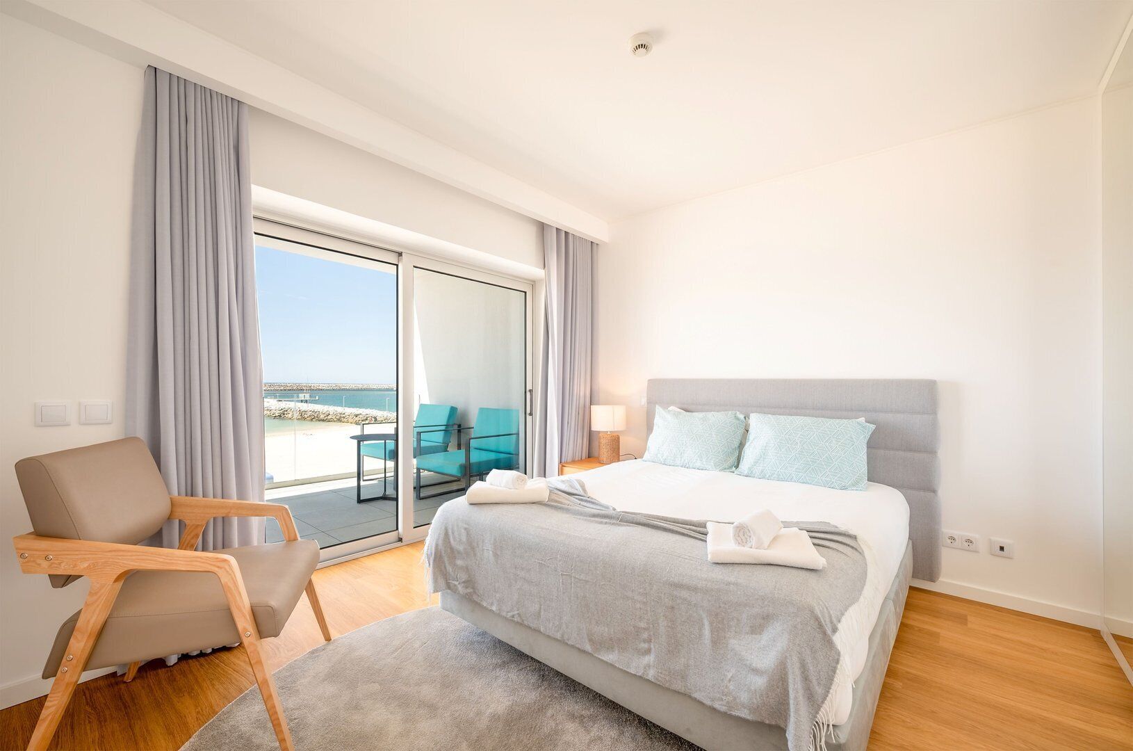 A modern hotel room with a large bed, a seating area with a chair, and a balcony with a view of the sea.