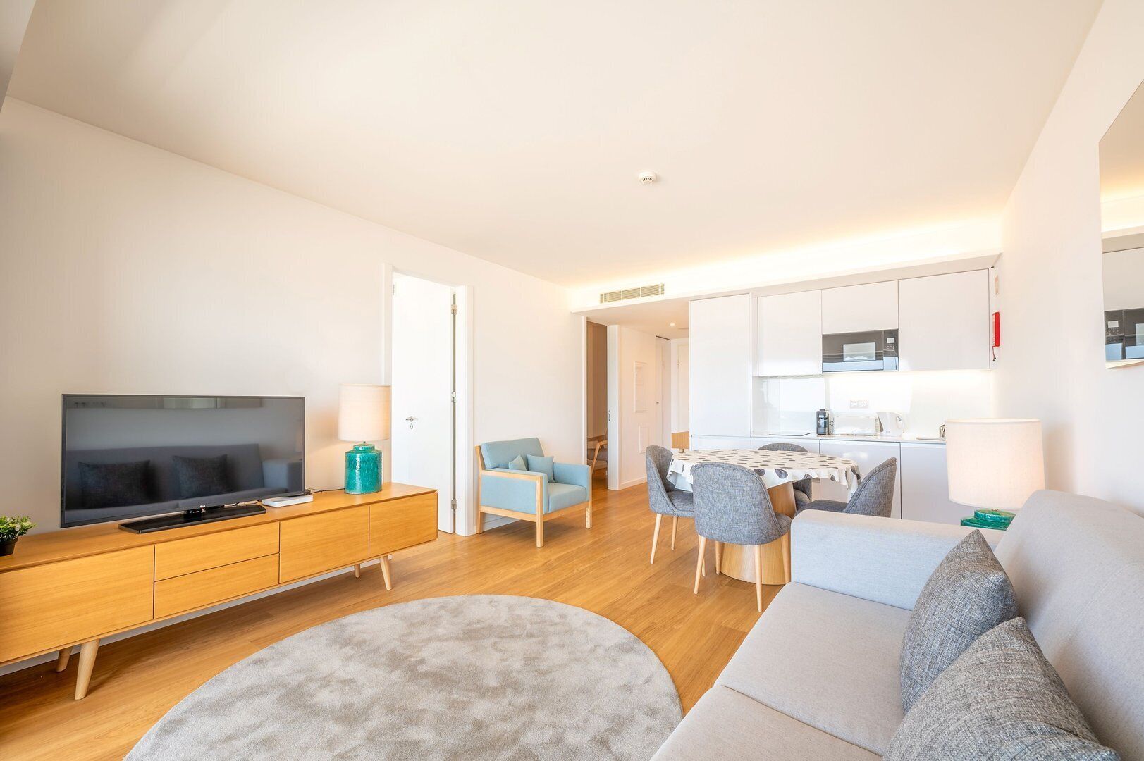 Un soggiorno moderno con una televisione a schermo piatto, un mobile porta TV in legno, un divano grigio, un tappeto rotondo e un'area pranzo con tavolo e sedie.