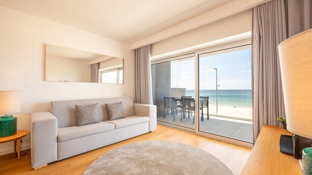 Ein modernes Wohnzimmer mit einer Couch, einem runden Teppich, einem Holztisch und Blick auf den Strand durch große Glasfenster.