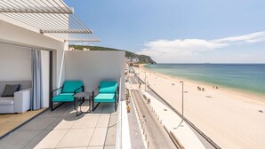 A balcony with two turquoise chairs and a small table, overlooking a beach with a clear view of the sea and distant buildings.