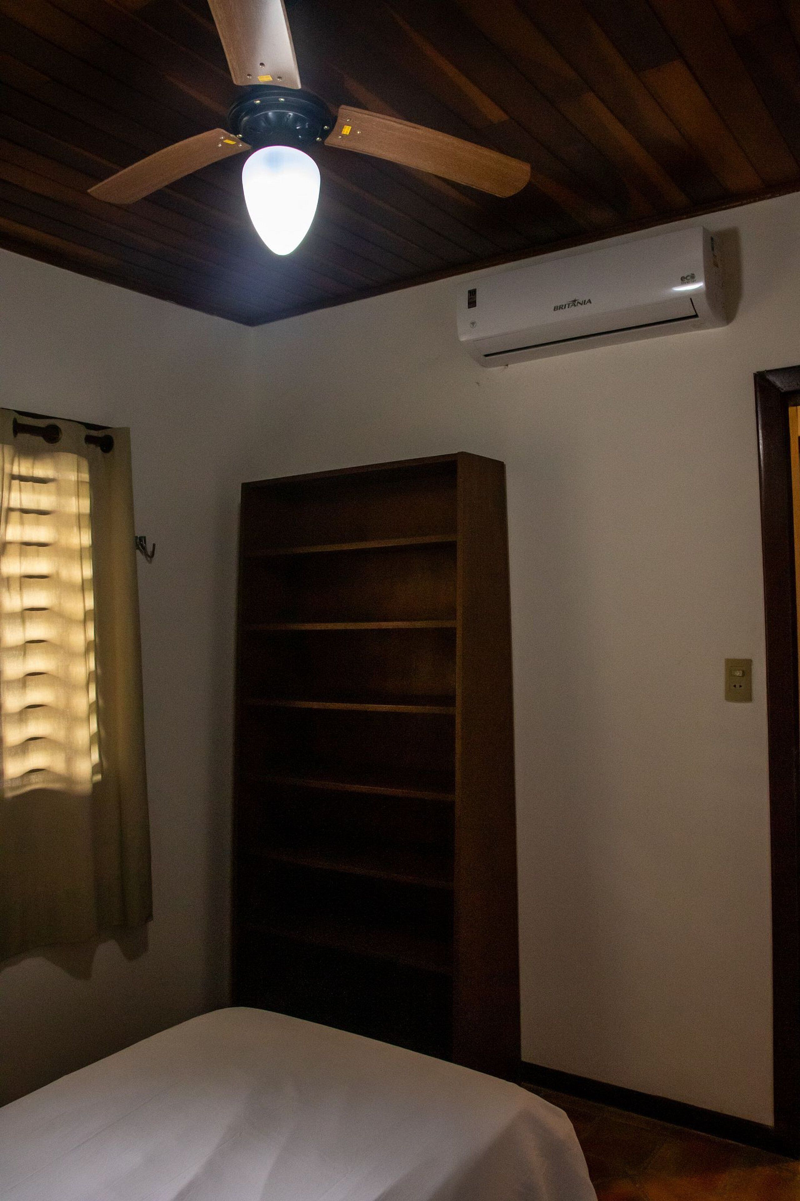 A bedroom with a bed, a ceiling fan, an air conditioning unit, a window with curtains, and a wooden wardrobe.