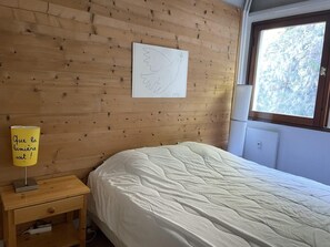 Un dormitorio con pared de madera, una cama con cubrecama blanco, una mesita de noche con lámpara y una ventana con vista a árboles.