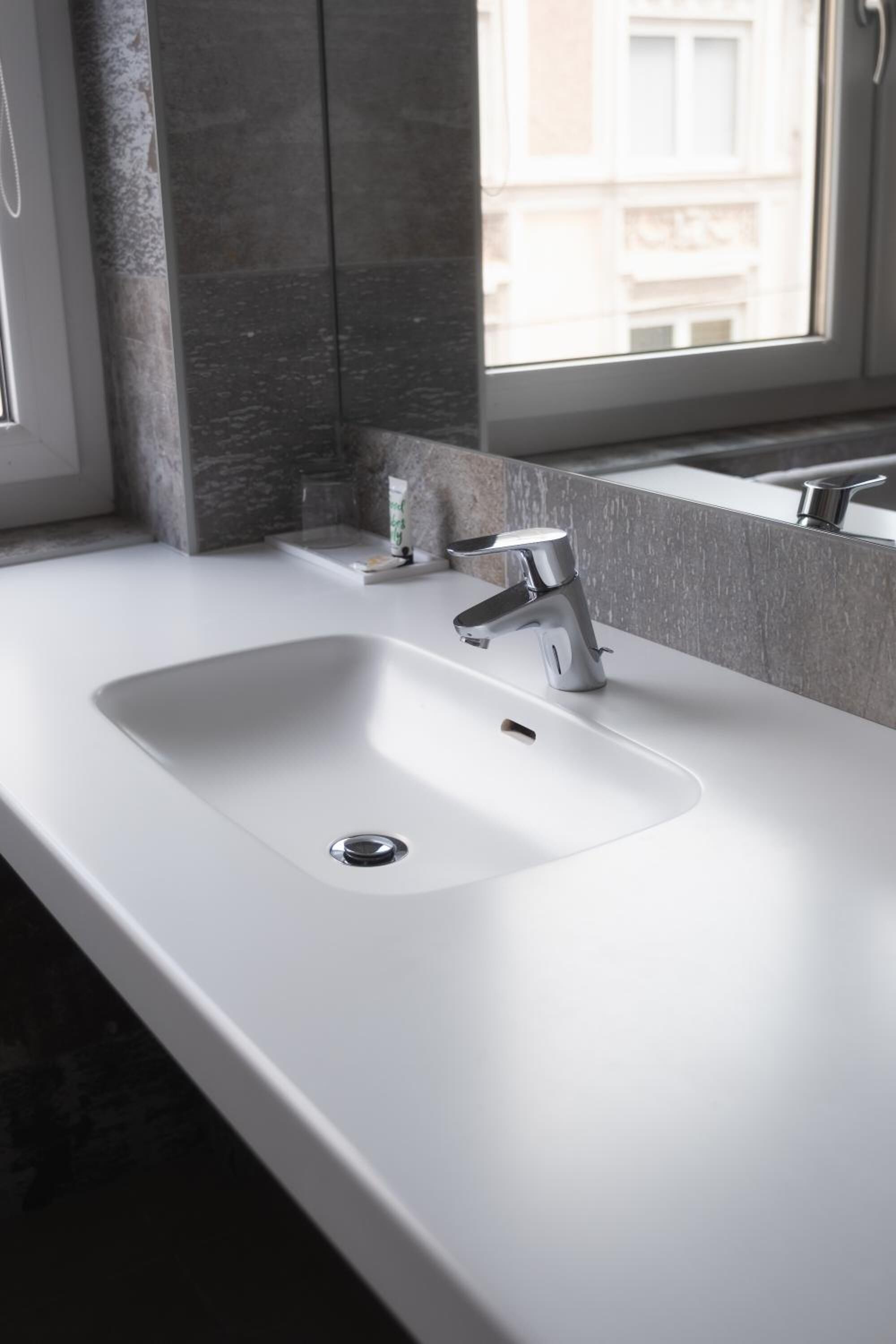 Un baño moderno con lavamanos blanco, grifo cromado y ventana con vistas a edificios.