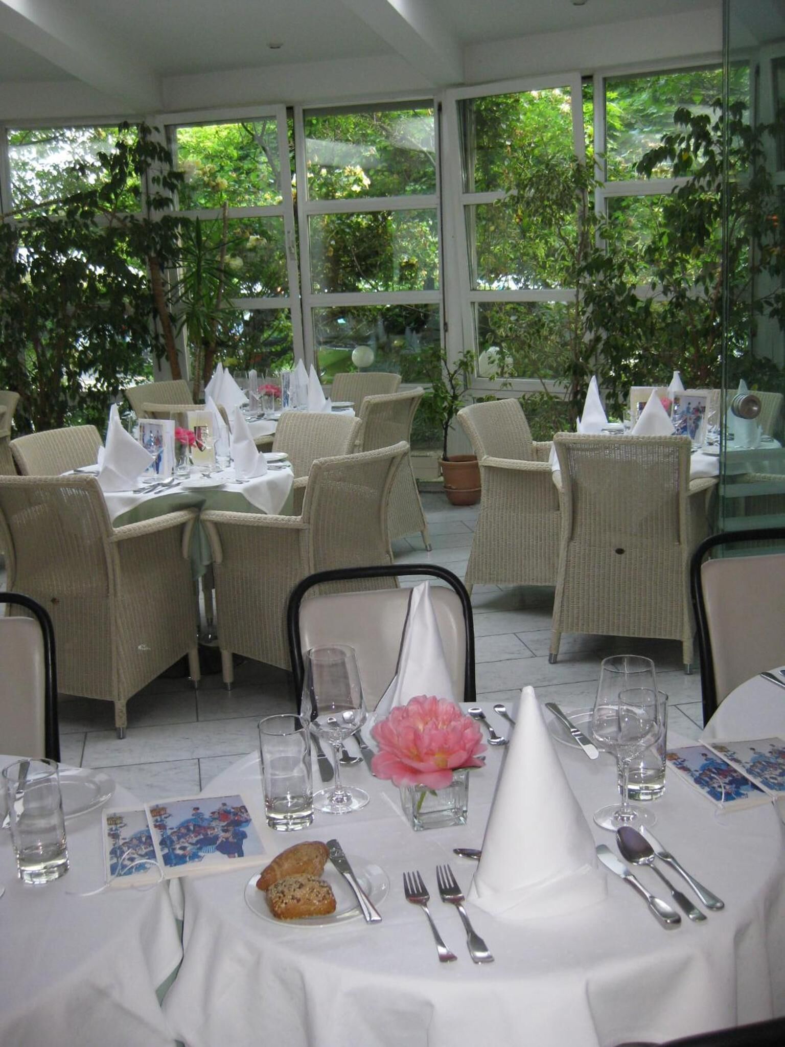 A restaurant with outdoor seating, white tablecloths, wicker chairs, and a view of greenery through large windows.