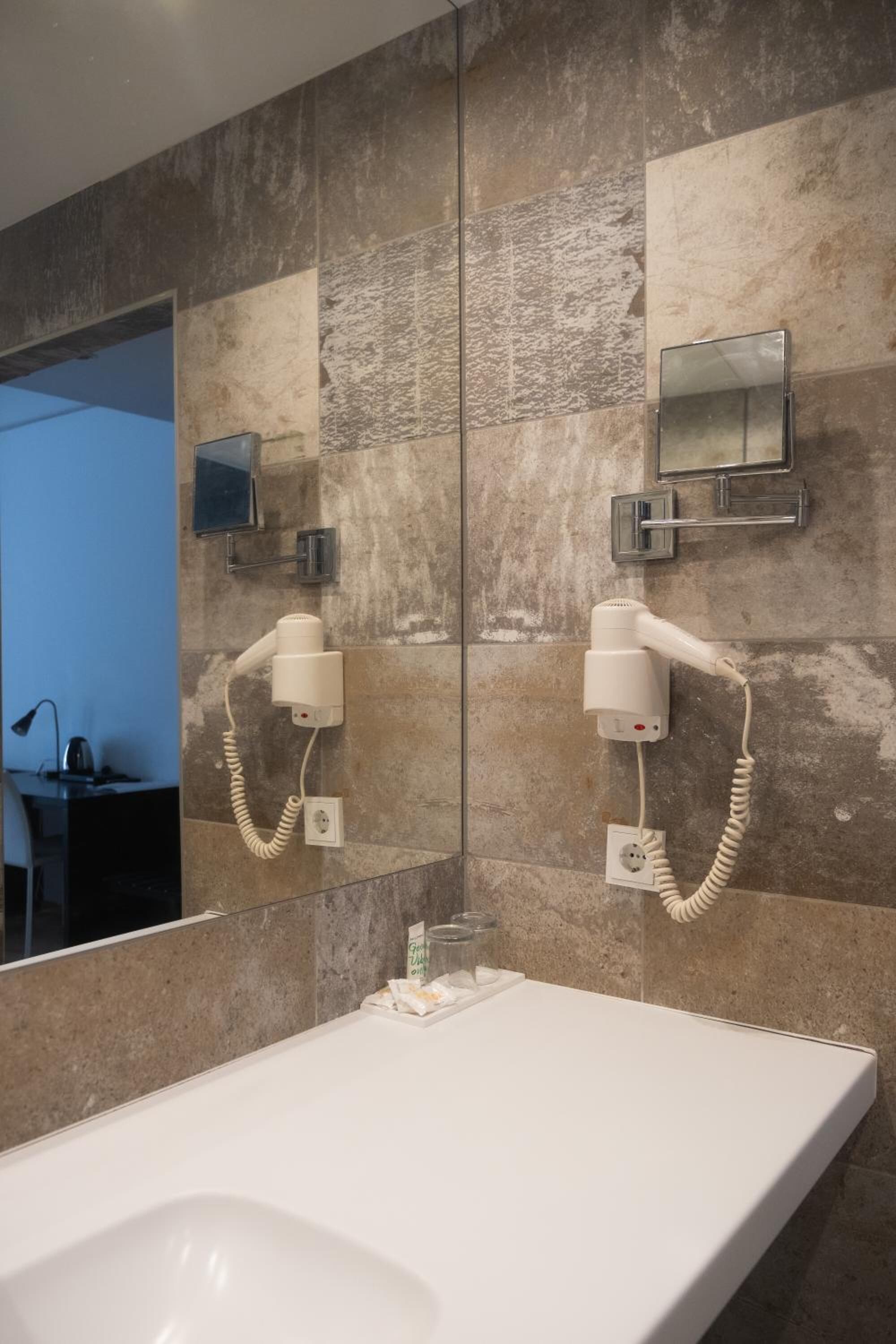 A modern bathroom with a large mirror, two hair dryers mounted on the wall, and a white sink.
