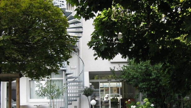 A modern building with a spiral staircase, a glass entrance, and a garden with various plants and a pathway. - City Hotel (Linz)