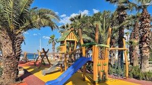 A playground with a blue slide and climbing structures, surrounded by palm trees and a clear sky. - Suelo Flat Villa for Family (Bodrum)
