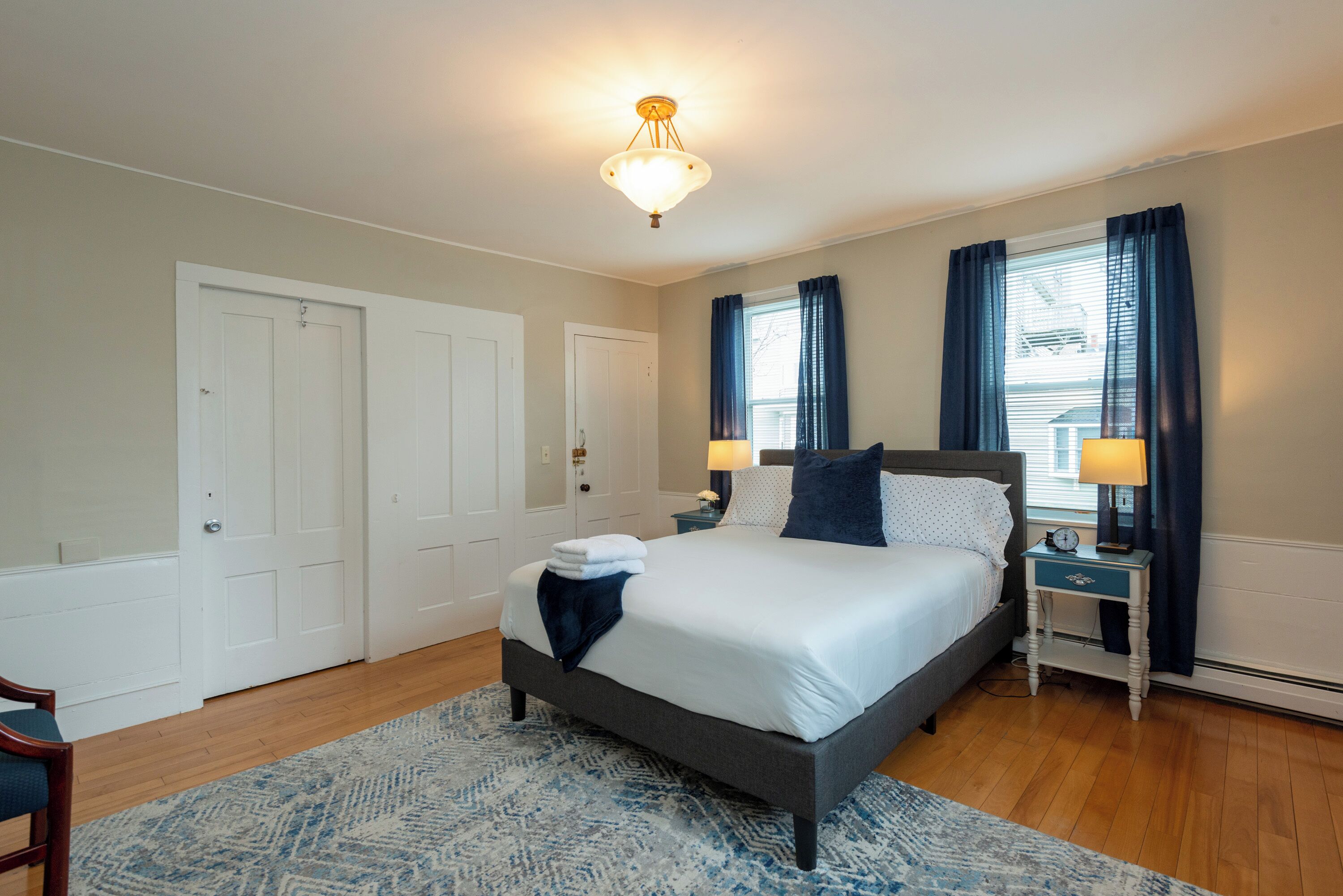 Une chambre à coucher comprenant un lit, deux tables de chevet, une commode, une chaise et une fenêtre avec des rideaux.