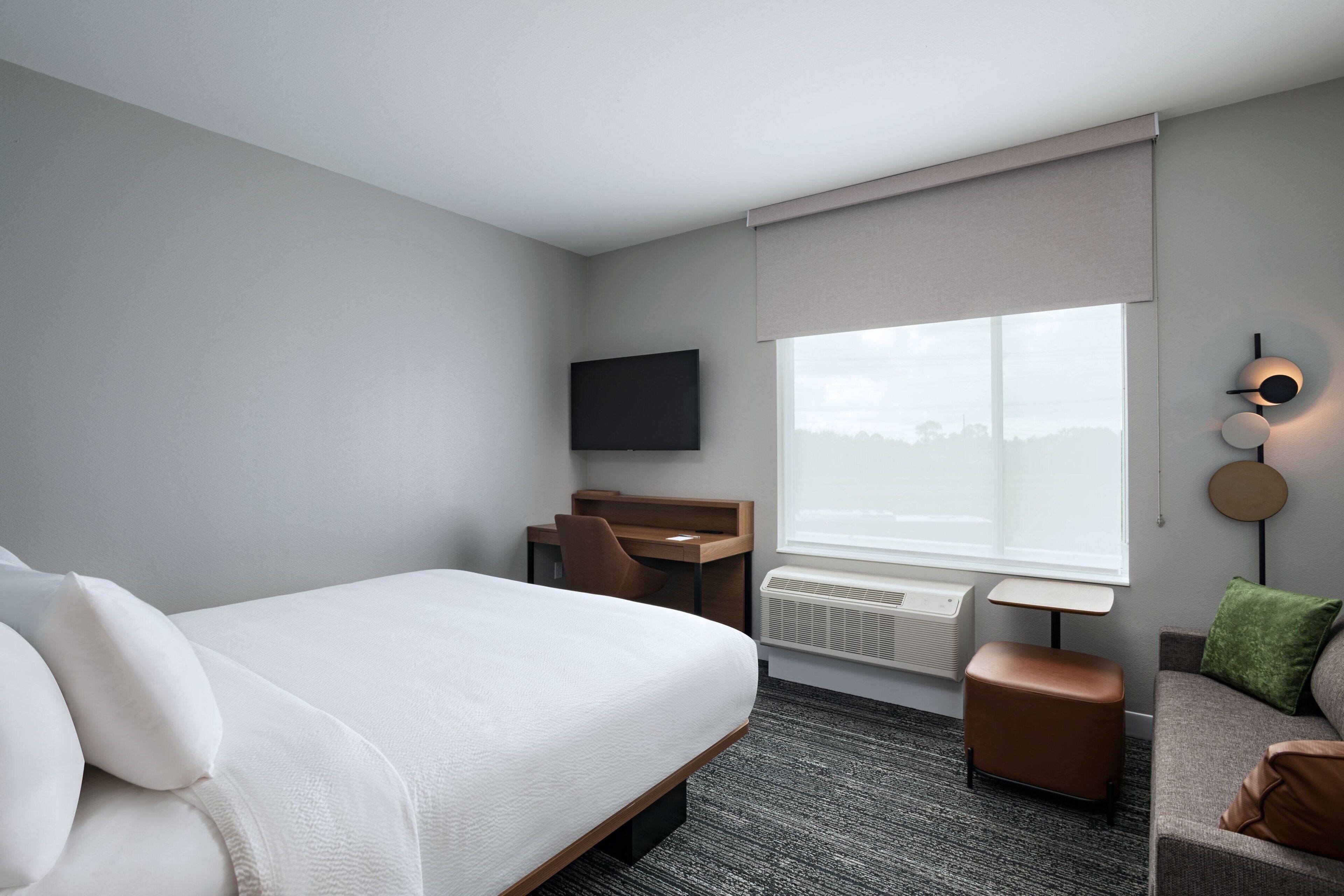 A modern hotel room with a large bed, a desk, a chair, a TV, a window with a view, and a small seating area with cushions.