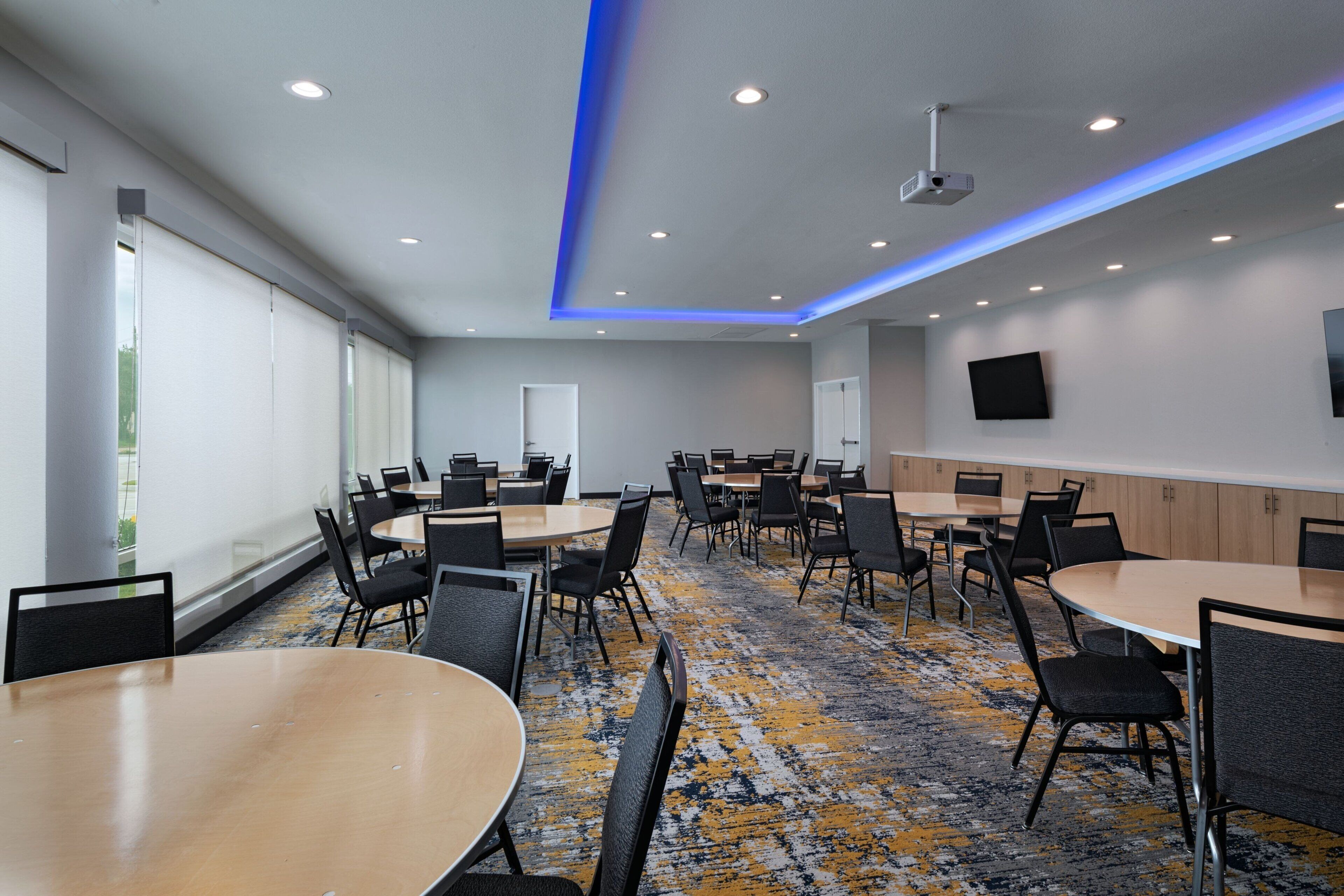 A conference room with multiple tables and chairs, a projector, and a large window with blinds.