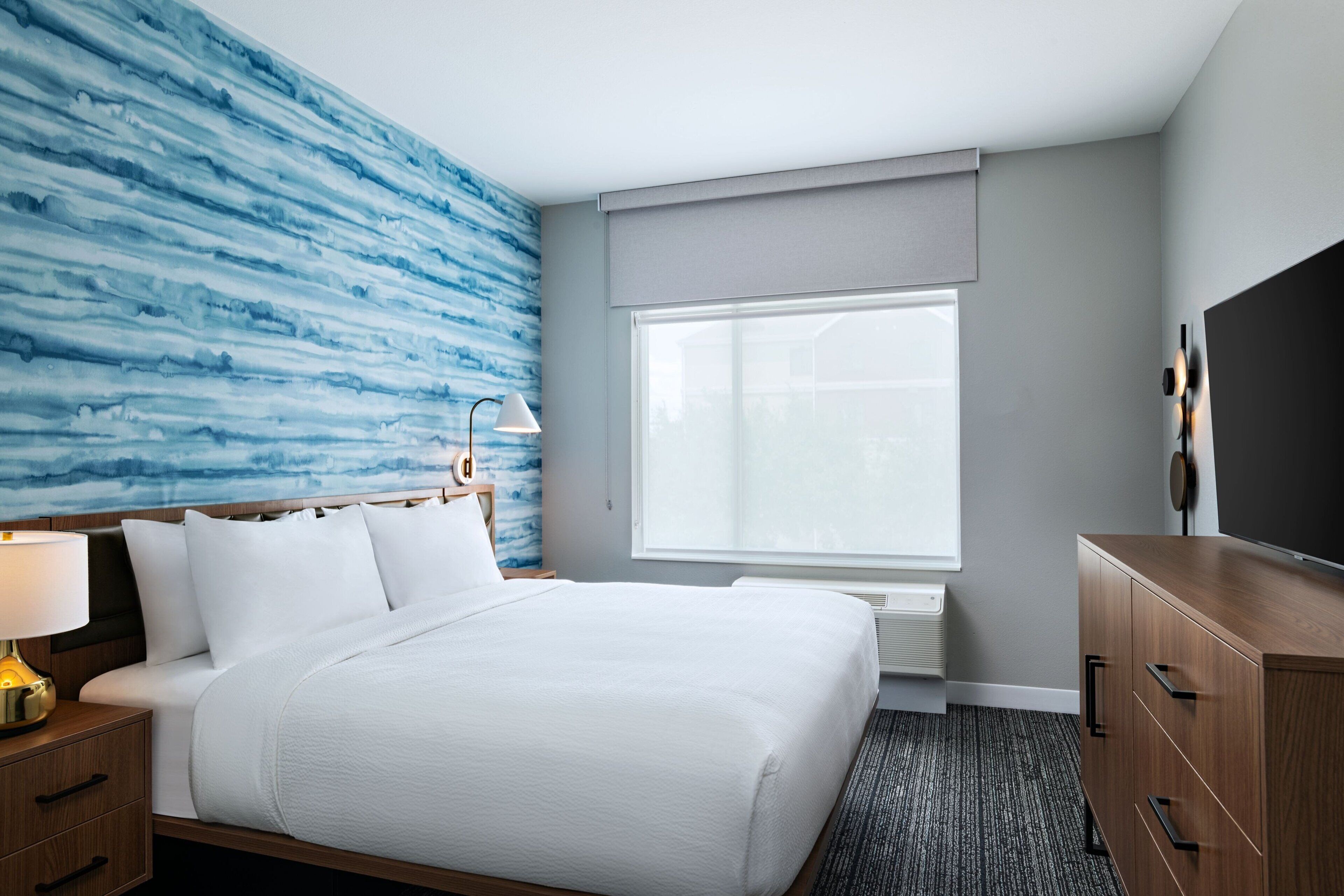Une chambre d’hôtel avec un lit, une table de chevet, une lampe, une commode et une grande fenêtre.
