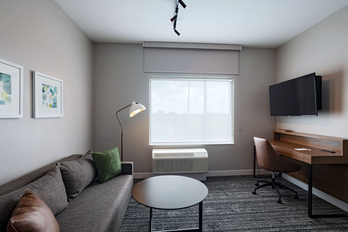 Une pièce moderne avec un canapé gris, une petite table ronde, un bureau avec une chaise, un téléviseur fixé au mur et une grande fenêtre avec des stores.