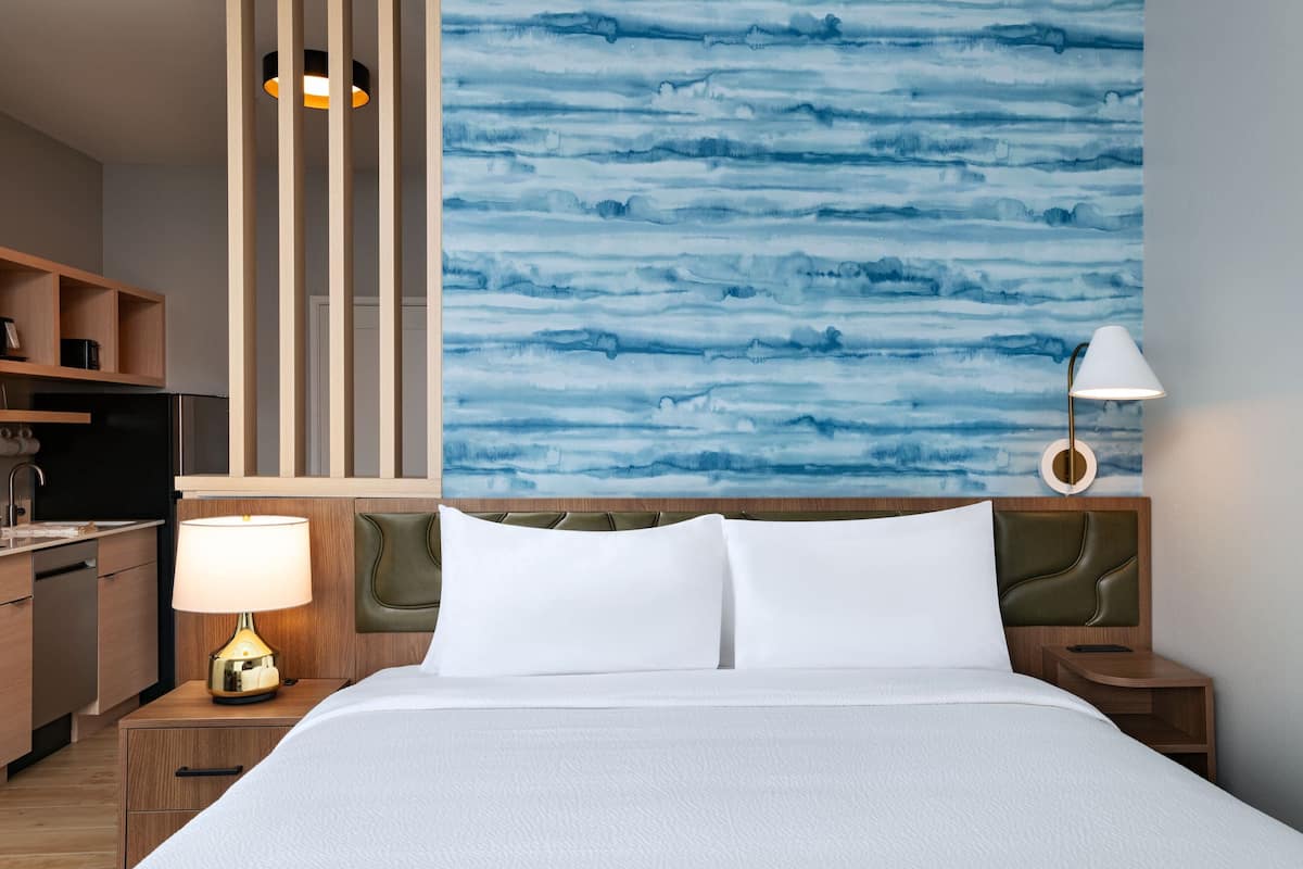 Une chambre d’hôtel comprenant un lit, une table de chevet avec une lampe, une tête de lit en bois, un mur orné d’un motif abstrait bleu et un coin cuisine.