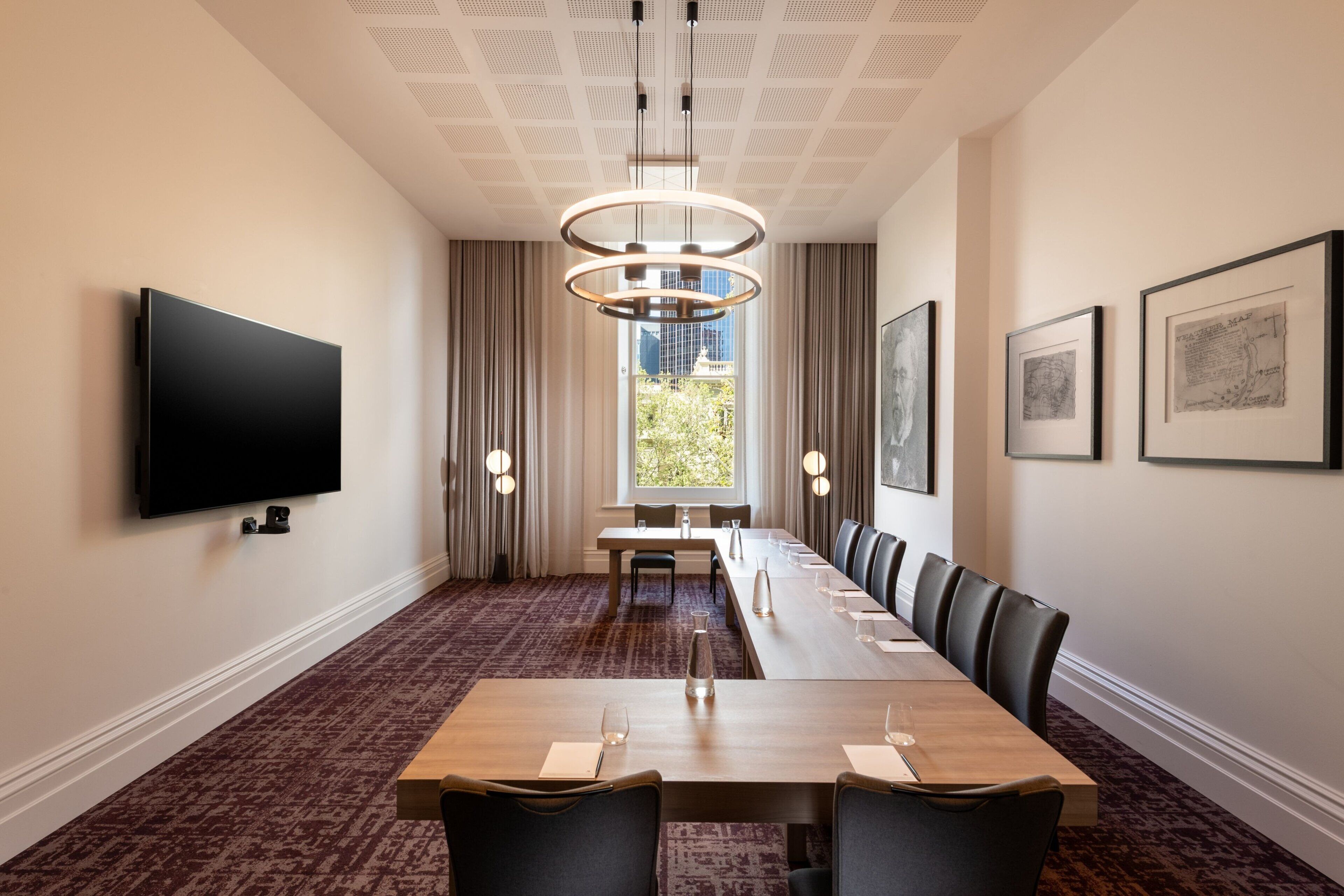 A conference room with a long table, chairs, a flat-screen TV, a chandelier, and framed artwork on the walls.