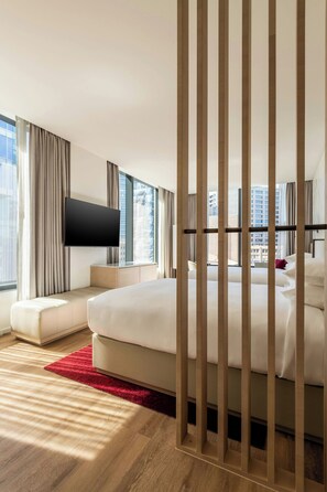 A modern hotel room with a large bed, a flat-screen TV mounted on the wall, and a view of the cityscape through large windows.