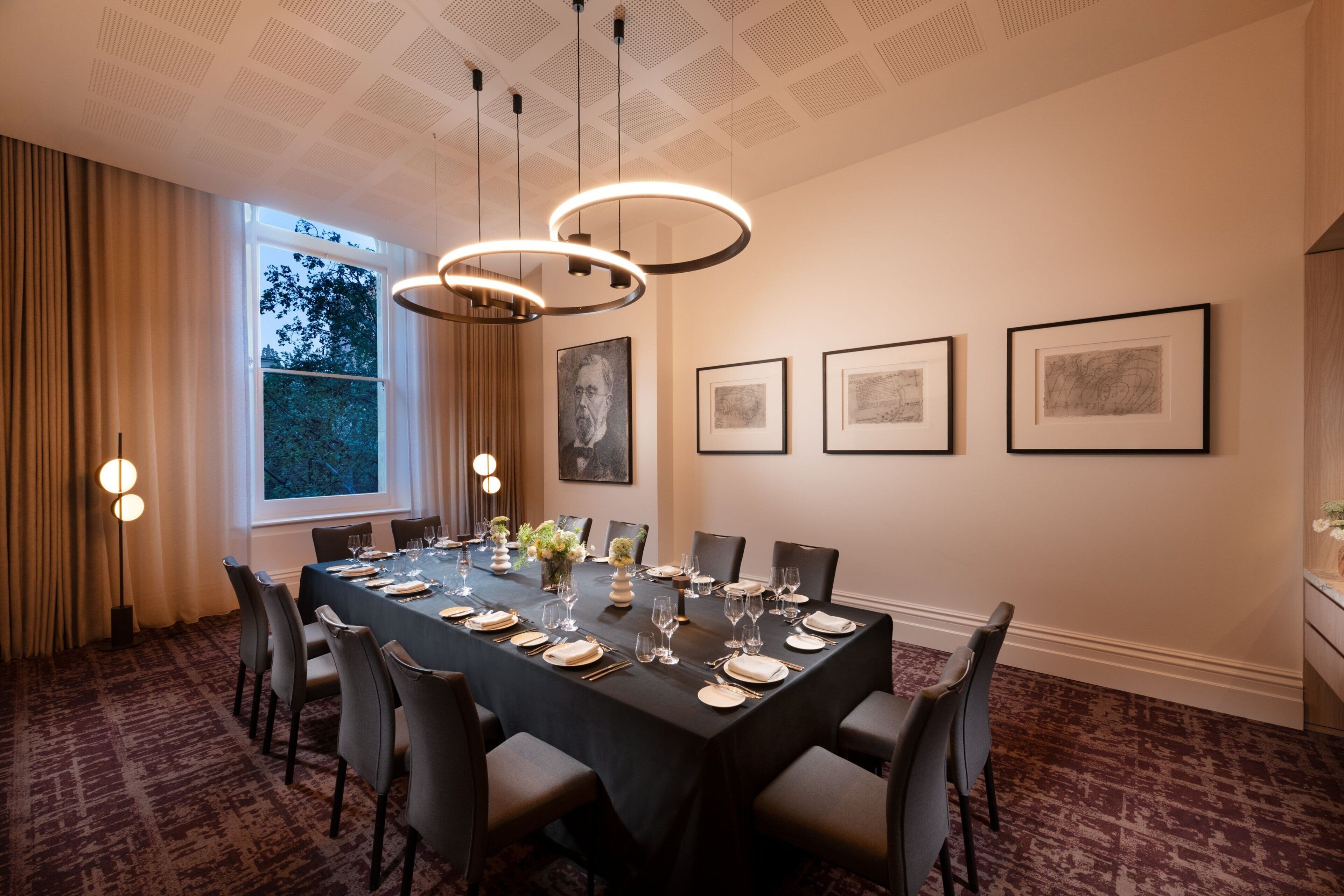 A dining room with a long table set for six, surrounded by chairs, with a large window and artwork on the wall.