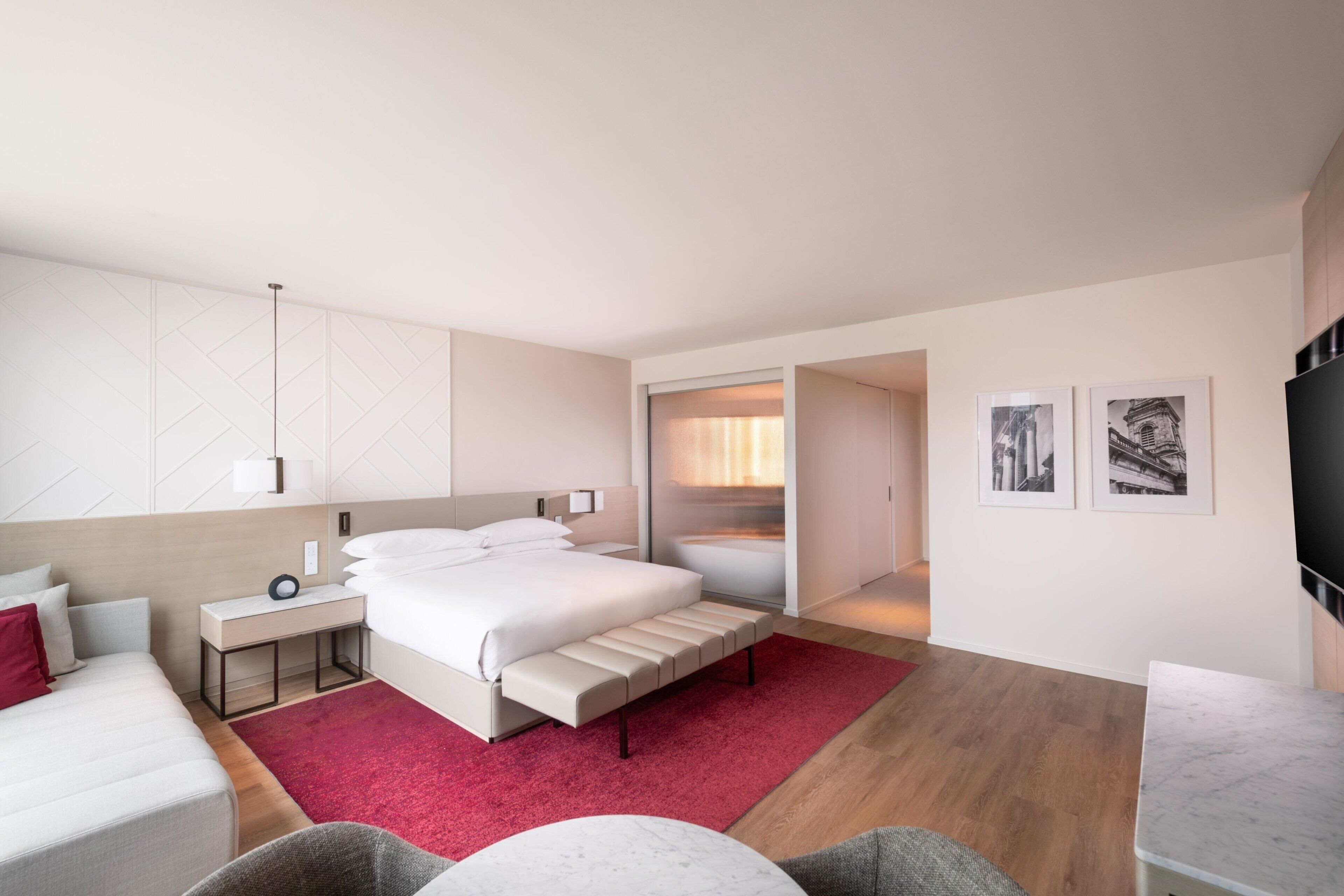 A modern hotel room with a large bed, a bedside table, a red rug, a seating area with a chair and a small table, and a TV on the wall.