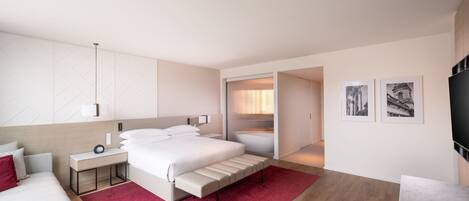 A modern hotel room with a large bed, a bedside table, a red rug, a seating area with a chair and a small table, and a TV on the wall.