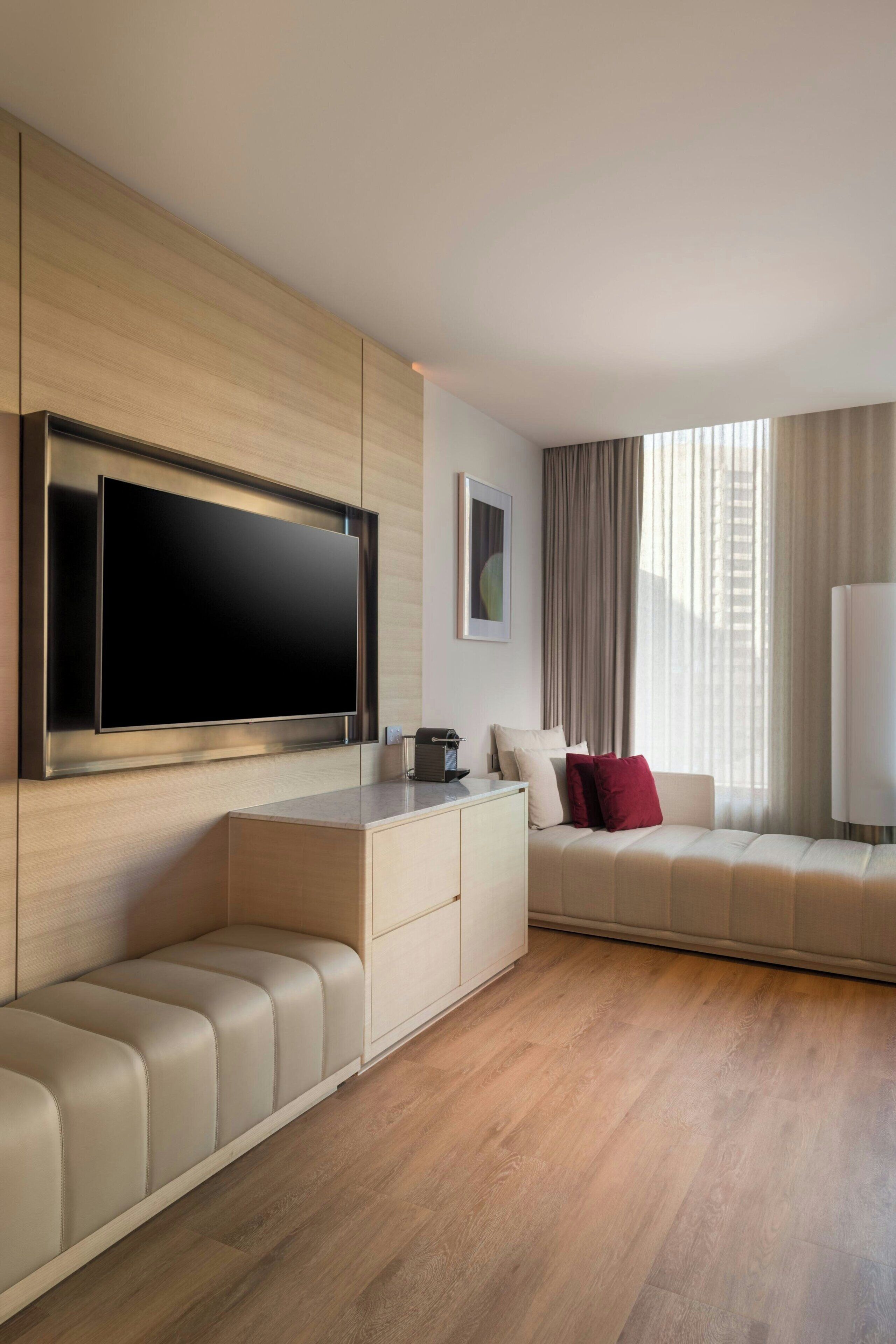 A modern living room with a large flat-screen TV mounted on a wooden wall, a beige sofa with red cushions, a low wooden bench, and a white side table.