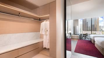 A modern hotel room with a marble bench, a walk-in closet with hanging robes, a bed with a red rug, a seating area with a table and chairs, and a city view through large windows.