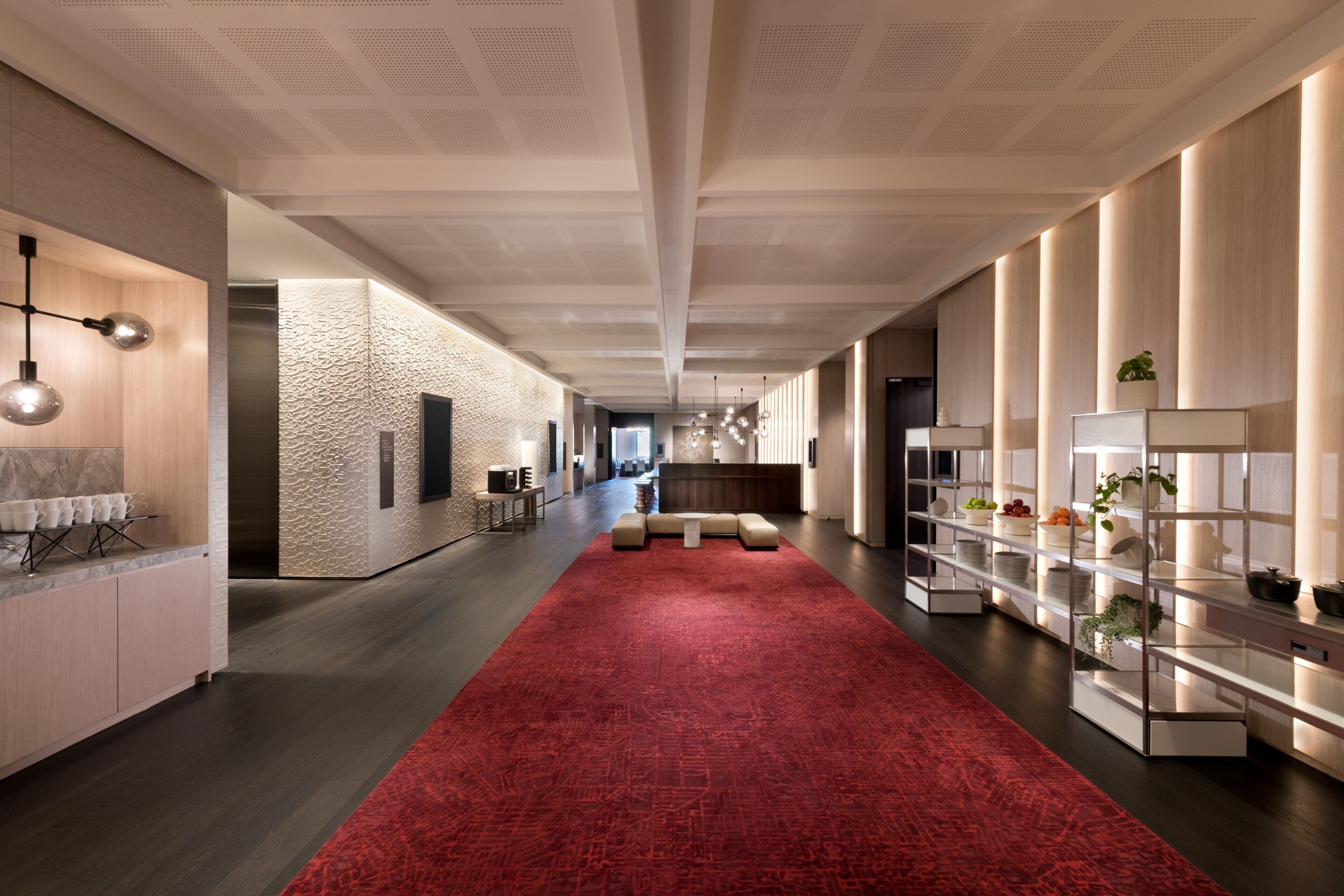 A modern hotel lobby with a red carpet, a reception desk, and a long hallway with a patterned wall and shelves with plants.