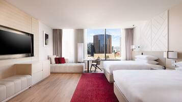 A hotel room with a large flat-screen TV, a sofa, two beds, a small table, and a view of the cityscape.