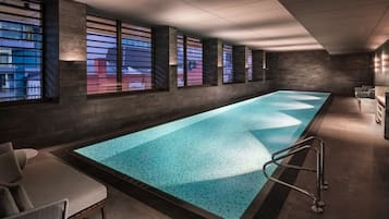 A modern indoor swimming pool with a long, rectangular shape, surrounded by dark stone walls and large windows with blinds.