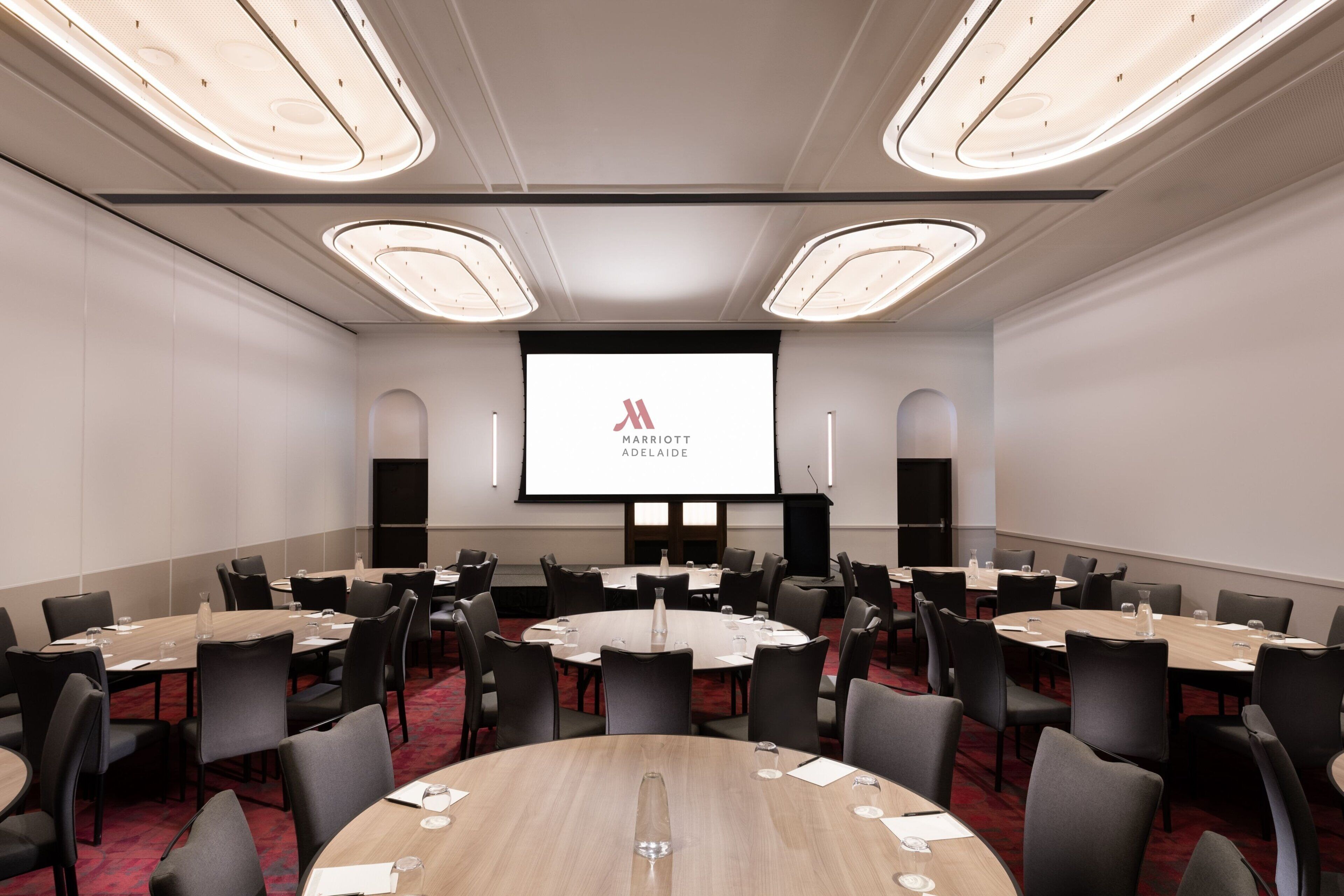 A conference room with round tables, black chairs, and a large screen displaying the Marriott logo.