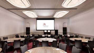 Een vergaderzaal met ronde tafels, zwarte stoelen en een groot scherm waarop het Marriott-logo staat.