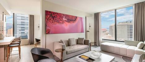Ein modernes Hotelzimmer mit großem Fenster, einer Couch, einem Couchtisch und einem Essbereich mit Stühlen.