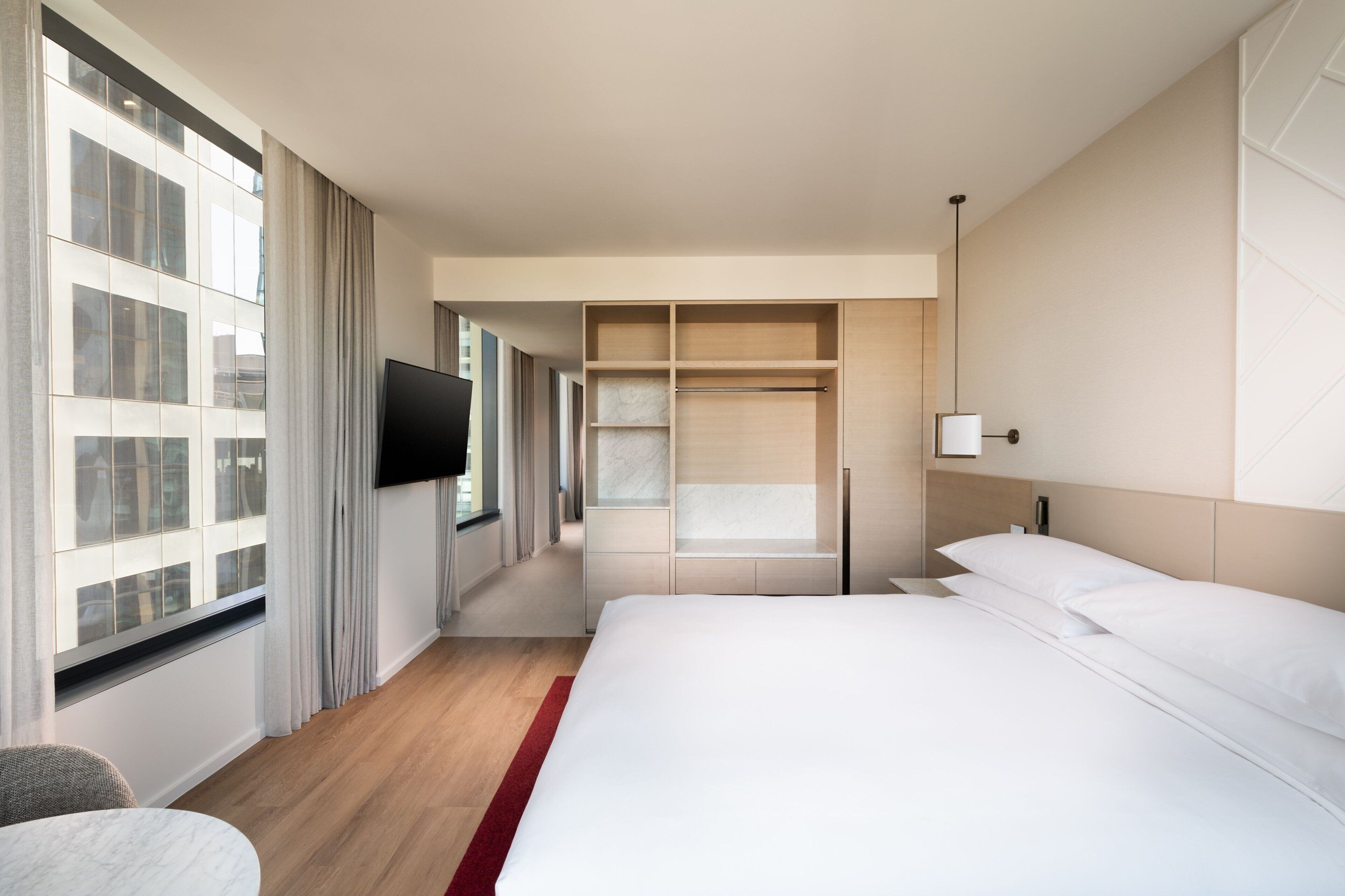 A hotel room with a large bed, a TV mounted on the wall, a walk-in closet, and a view of the city through large windows.