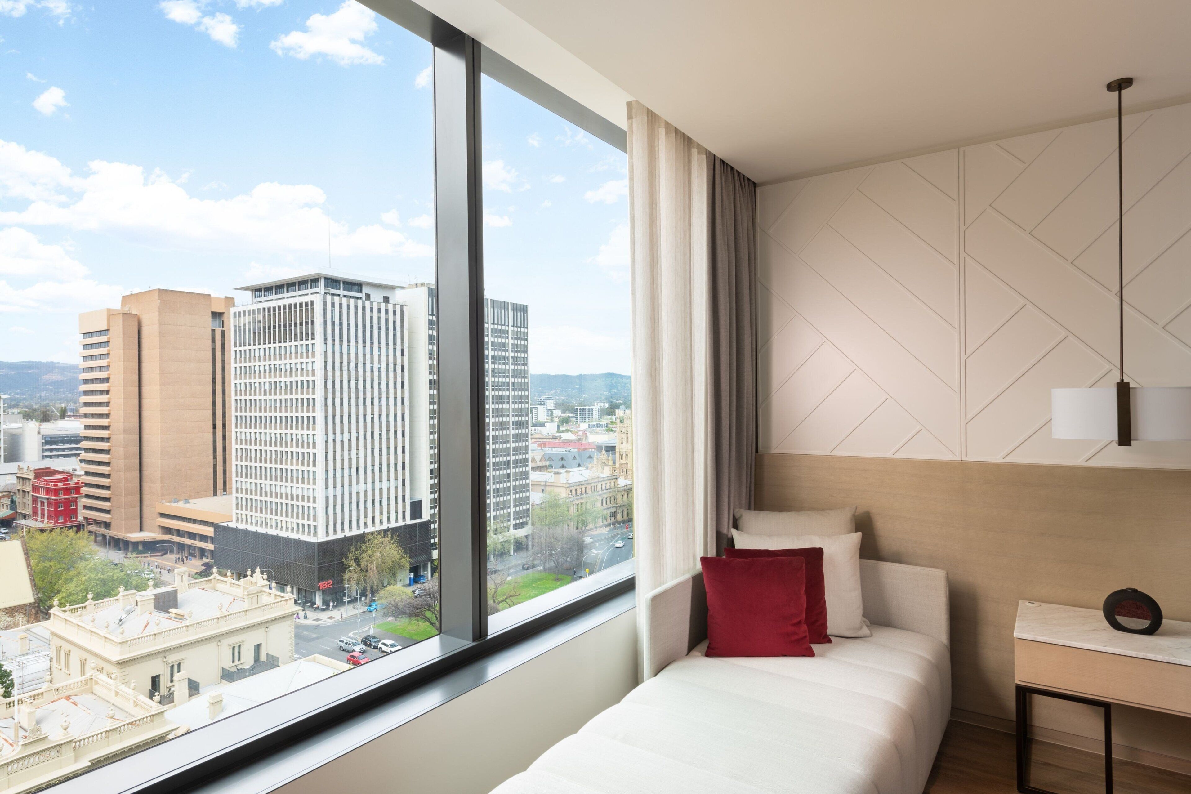 A modern hotel room with a large window offering a city view, a neatly made bed, a bedside table, and a hanging light fixture.